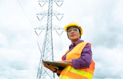 Electrical engineer woman holds laptop and stands in front of electric power transmission lines.
